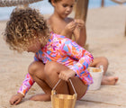 Girl playing with sand on the beach wearing Snapper Rock One Piece UPF 50+ Swimsuit - Blooming Sunset. Available at www.tenlittle.com