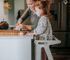 Child standing on All Circles Toddler Tower in white in the kitchen while helping adult with cooking. Available from tenlittle.com