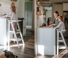 Two views of child standing on All Circles Toddler Tower in white in the kitchen - one view of child by themself, one view of child with adult. Available from tenlittle.com