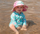 Child wearing Jan & Jul Long Sleeve UV Sun Suit in Watermelon while sitting in the water, wearing a bucket hat. Available from tenlittle.com