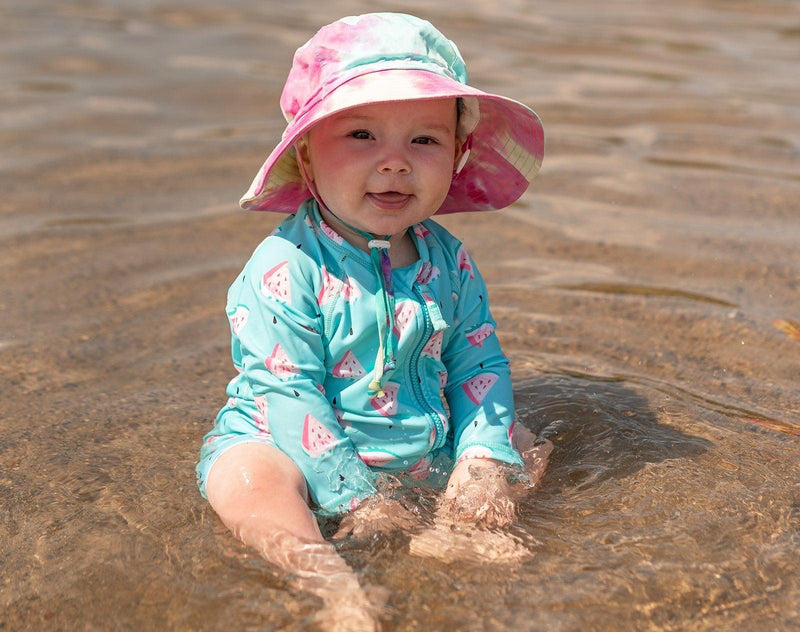 Child wearing Jan & Jul Long Sleeve UV Sun Suit in Watermelon while sitting in the water, wearing a bucket hat. Available from tenlittle.com