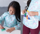 Girl drawing and putting markers in her Bumkins Rainbow Long Sleeved Bib. Available from www.tenlittle.com.