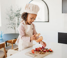 Child cutting fruits in the Piccalio Mini Chef Apron & Hat Set in beige. Available from www.tenlittle.com