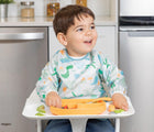 Child eating in highchair wearing Bumkins Sleeved Bib in dinosaurs. Available at tenlittle.com