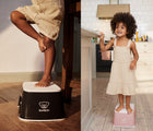 One view of child standing on BabyBjörn Step Stool in black, and another view of child in kitchen standing on BabyBjörn Step Stool in pink. Available from tenlittle.com