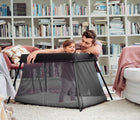 Child standing inside of BabyBjörn Travel Crib Light in black with adult nearby looking at child, in a living room with a bookshelf. Available from tenlittle.com