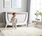 Child standing next to bassinet crib in a bedroom, next to the window, from Béaba Air Complete Sleep System Bassinet-to-Crib. Available from tenlittle.com