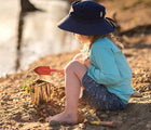 Child wearing Jan & Jul UV Swim Shorts in Shark with UV Long Sleeve Top in lagoon and navy bucket hat, outside digging in the dirt. Available from tenlittle.com