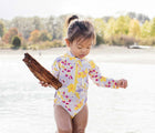 Girl wearing Jan & Jul Long One Piece UV Swimsuit in Tropical Bloom near the water and trees, and holding a piece of wood. Available from tenlittle.com