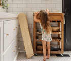 Child standing next to Piccalio Foldable Kitchen Tower in natural which is folded next to the cabinet. Available from tenlittle.com
