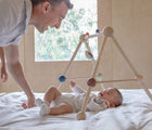 Baby laying underneath PlanToys Wooden Play Gym in orchard on a bed with adult nearby smiling. Available from tenlittle.com
