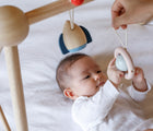 Baby on bed holding one of the hanging toys from PlanToys Wooden Play Gym in orchard with adult hand helping. Available from tenlittle.com