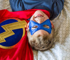 Child wearing blue side of mask from Little Adventures Hero Cape and Mask costume and laying down on bed. Available from tenlittle.com