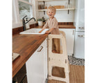 Boy standing on Piccalio Convertible Kitchen Helper Tower in natural in the kitchen next to the sink. Available from tenlittle.com