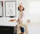 Girl wearing Piccalio Mini Chef Apron & Hat Set in beige in the kitchen while standing on convertible kitchen tower. Available from tenlittle.com